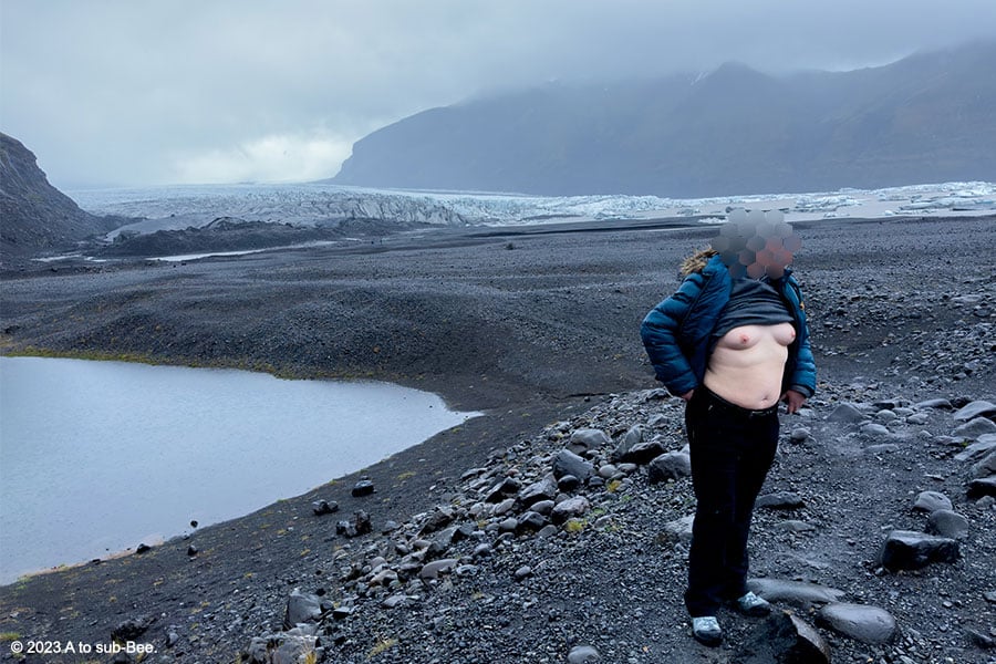 Bee standing on a black ash field with a glacier in the background baring their breasts.