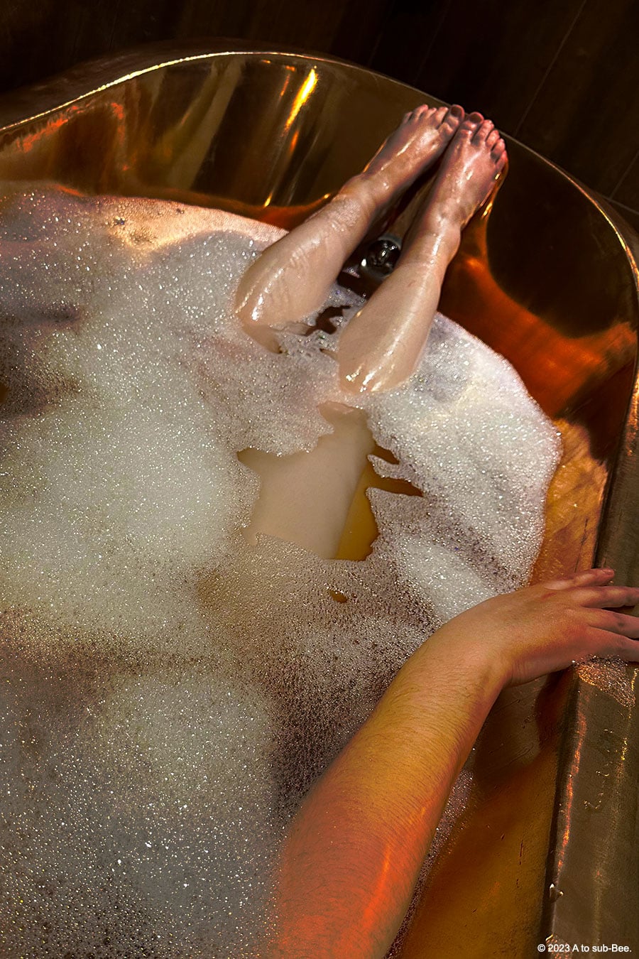 A person laying naked in a copper roll top bath covered in bubbles, pampering themselves