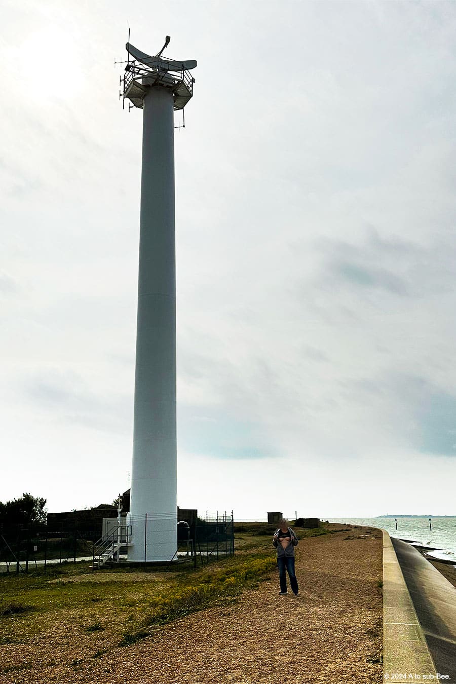 A tall radar tower with a person baring theor breasts in front of it, against the back drop of a coastline on an overcast day