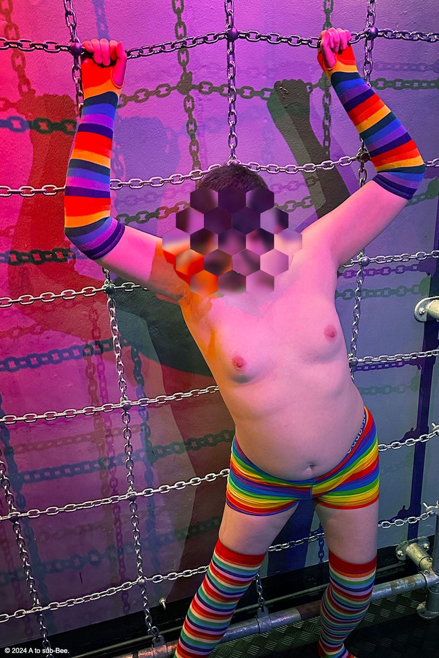 A person stood in front of a wall of chains wearing long rainbow socks, long rainbow gloves and rainbow boxers