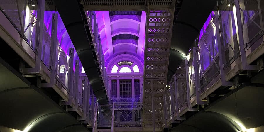 An image of a person standing on the second floor of an old prison cell block, the photographer is on the ground floor looking up at them and the lihting in the ceiling is purple