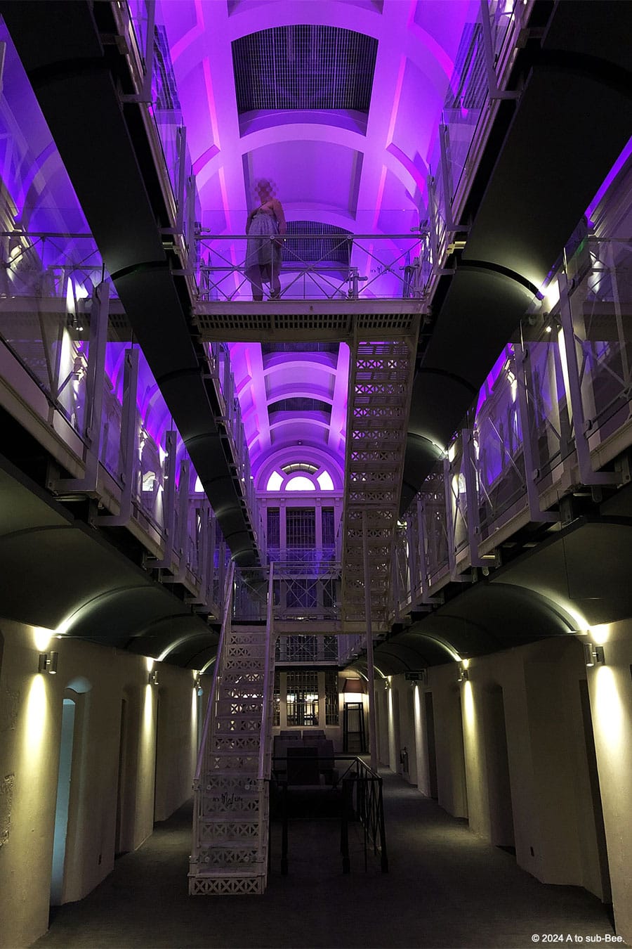 An image of a person standing on the second floor of an old prison cell block, the photographer is on the ground floor looking up at them and the lihting in the ceiling is purple