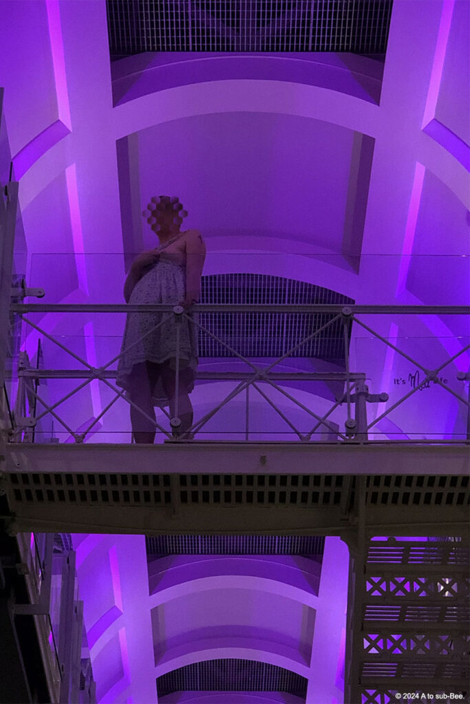 An closer image of a person standing on the second floor of an old prison cell block, bearing their breasts, the photographer is on the ground floor looking up at them and the lihting in the ceiling is purple