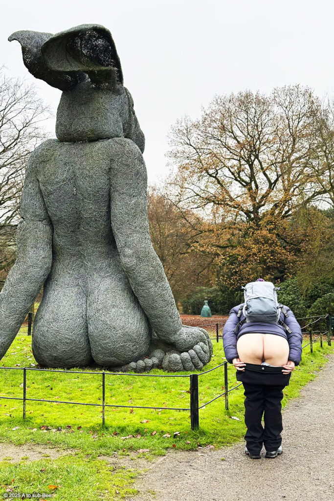 A person stood slightly bent over beside the back of a scuplture of a rabbit so both bottoms are side by side.