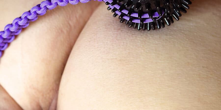 Image of a persons arse with a glimpse of their cunt between their legs. Balanced on their bottom in a black spikey ball with a purple paracord handle